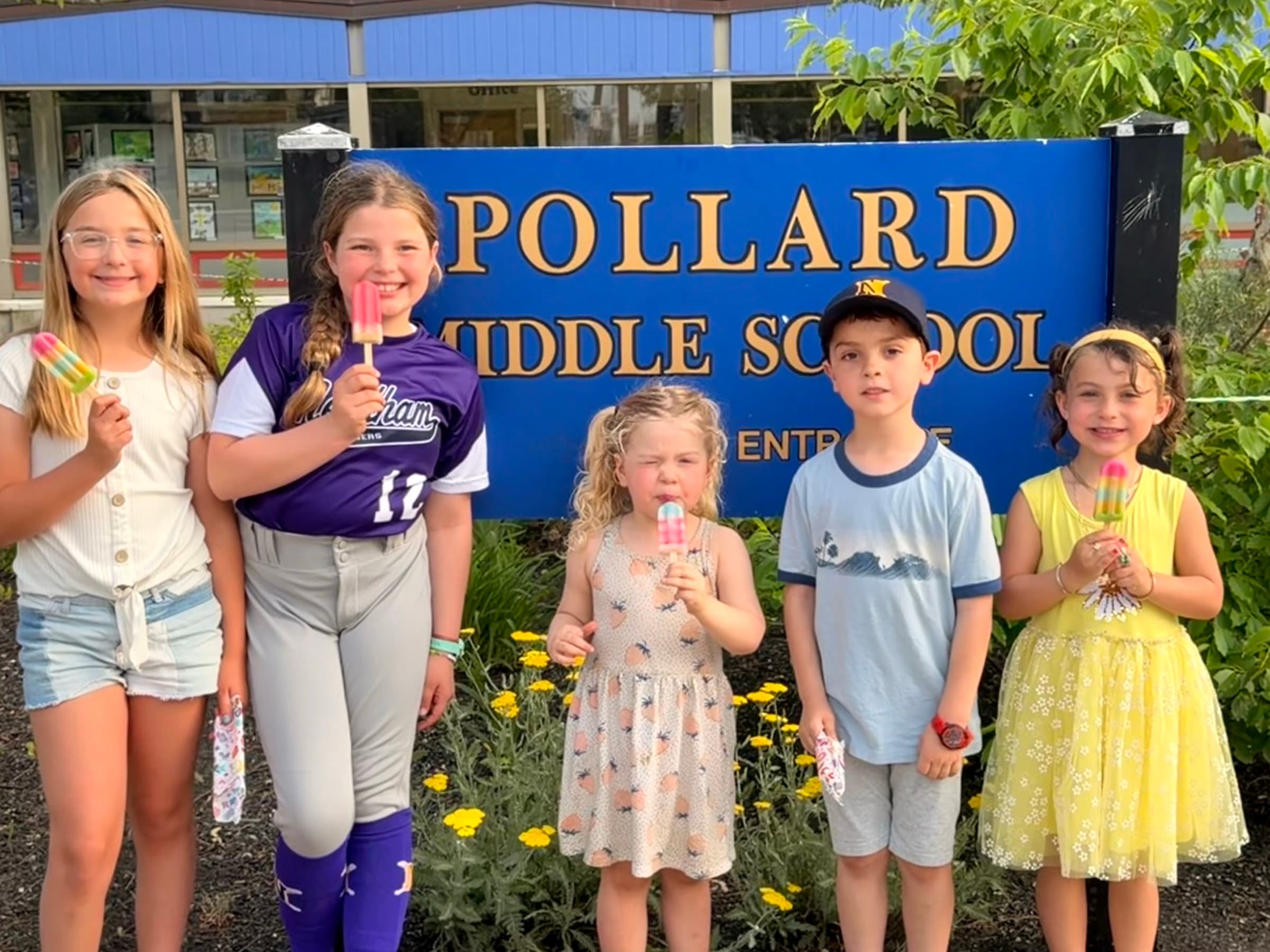 Five smiling kids holding popsicles in front of the blue Pollard Middle School entrance sign.